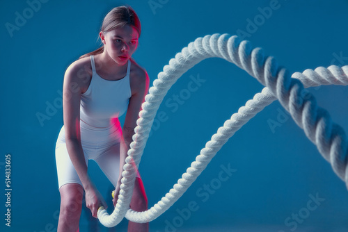 Strong athletic girl doing sports exercises with combat ropes while fitness workout routine. Long exposure, motion blur