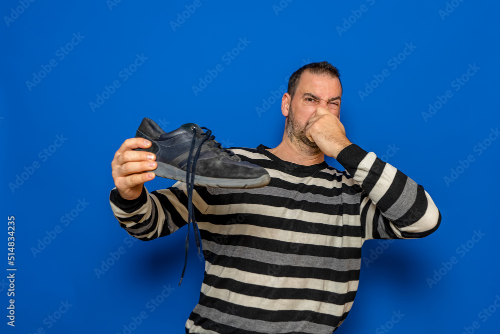 Naklejka premium Hispanic man with a beard dressed in a striped sweater holding his nose because of the bad smell that his sneaker gives off, isolated on blue studio background.