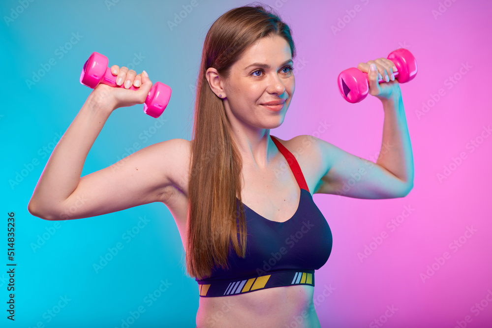 smiling sporty woman in sports bra doing exercise with pink dumbbells looking away. Female fitness portrait isolated on neon multicolor background. smiling sporty woman in sports bra doing exercise with pink dumbbells looking away. Female fitness portrait isolated on neon multicolor background.