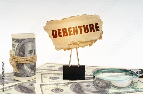 Among the dollars lies a magnifying glass and there is a clip and a cardboard sign with the inscription - Debenture