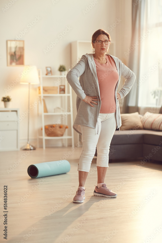 Portrait of serious senior woman in sportswear and glasses standing in living room and holding hands on hips