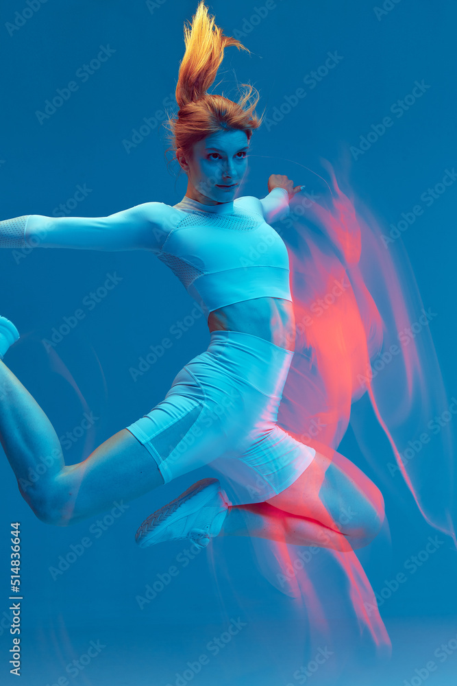 Jumping sports girl in air on blue backdrop. Colorful neon light, long exposure. Motion blur. Sport lifestyle, wellness