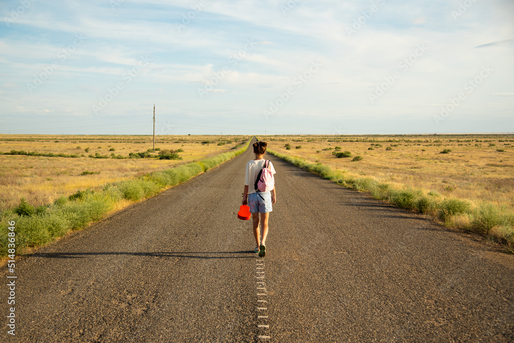 Obraz premium A girl walks along an empty road in the steppe. She has a ukulele in her hands and a backpack on her shoulders. It's summer and warm outside. Open road.
