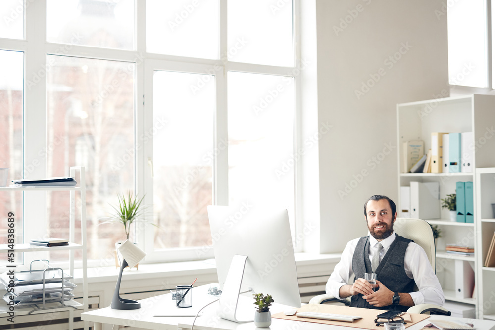 Portrait of cheerful successful young bearded businessman with beard sitting at table in spacious office full of light and drinking water from glass Portrait of cheerful successful young bearded businessman with beard sitting at table in spacious office full of light and drinking water from glass