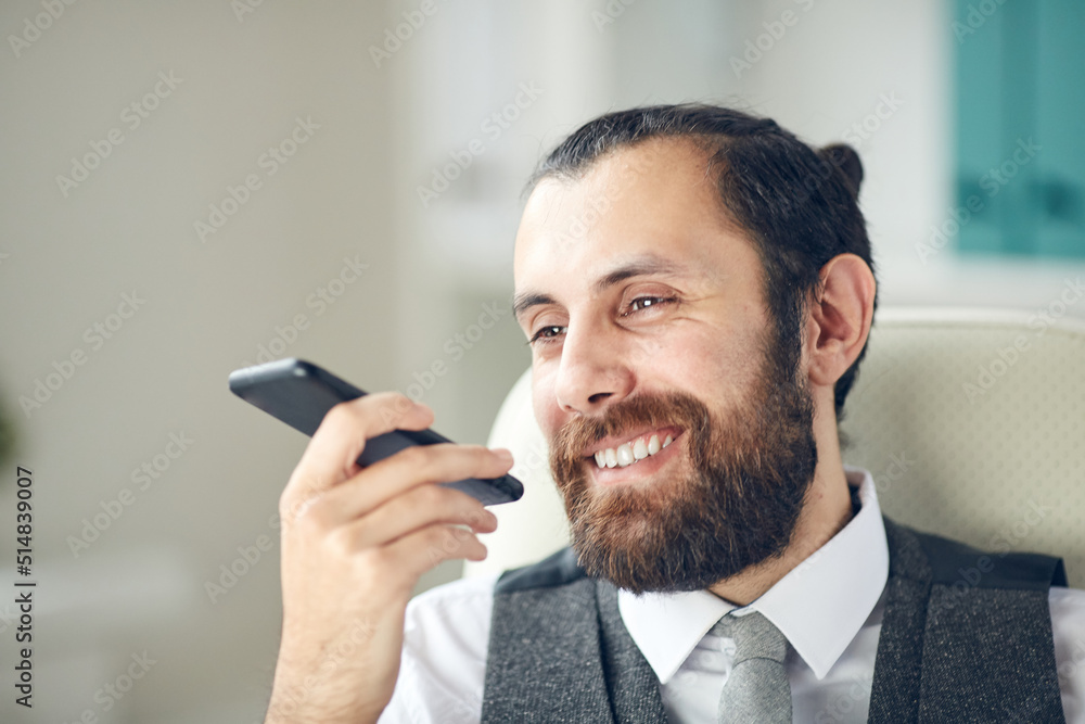 Positive handsome bearded businessman sitting on office chair and using smartphone while recording funny message for colleague Positive handsome bearded businessman sitting on office chair and using smartphone while recording funny message for colleague