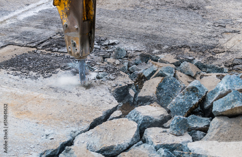 Close-up of demolition hammer with dust and sparks and broken concrete pieces. Detail of hydraulic jackhammer mounted on excavator arm working at old road reconstruction. Repairs in civil engineering.