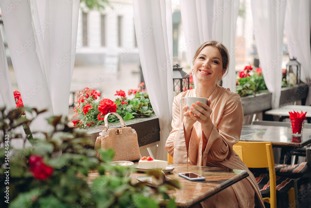A woman in a dress is sitting at a table in a cafe, drinking coffee and smiling, there is a phone nearby