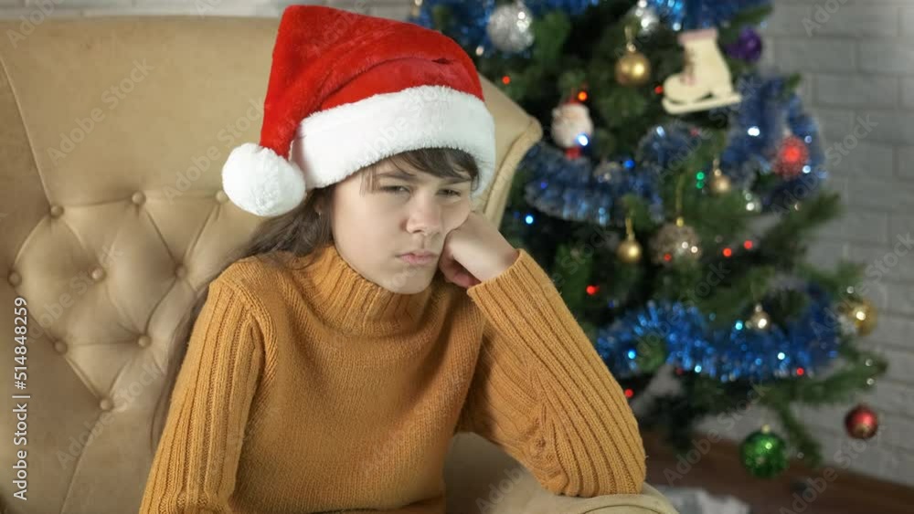 Unhappy teen by Christmas tree. A sad teen girl in Santa hat sits in loneliness in armchair by Christmas tree. Unhappy teen by Christmas tree. A sad teen girl in Santa hat sits in loneliness in armchair by Christmas tree.