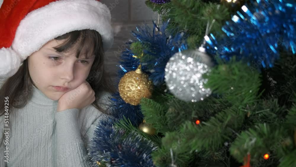 Child feeling sad at New Year eve. A little girl feeling sad in her Santa red hat by the winter decorated tree in the room. Child feeling sad at New Year eve. A little girl feeling sad in her Santa red hat by the winter decorated tree in the room.