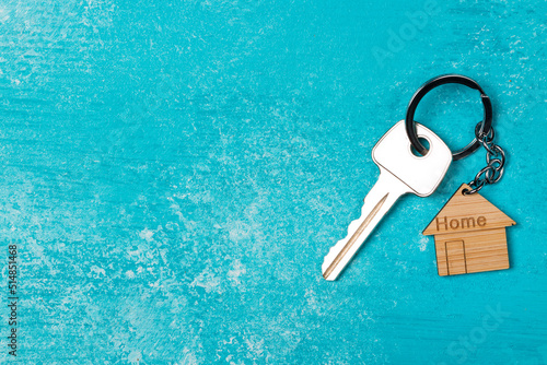Wooden house shaped keychain and key on blue wooden background. Free space © Nikolay N. Antonov