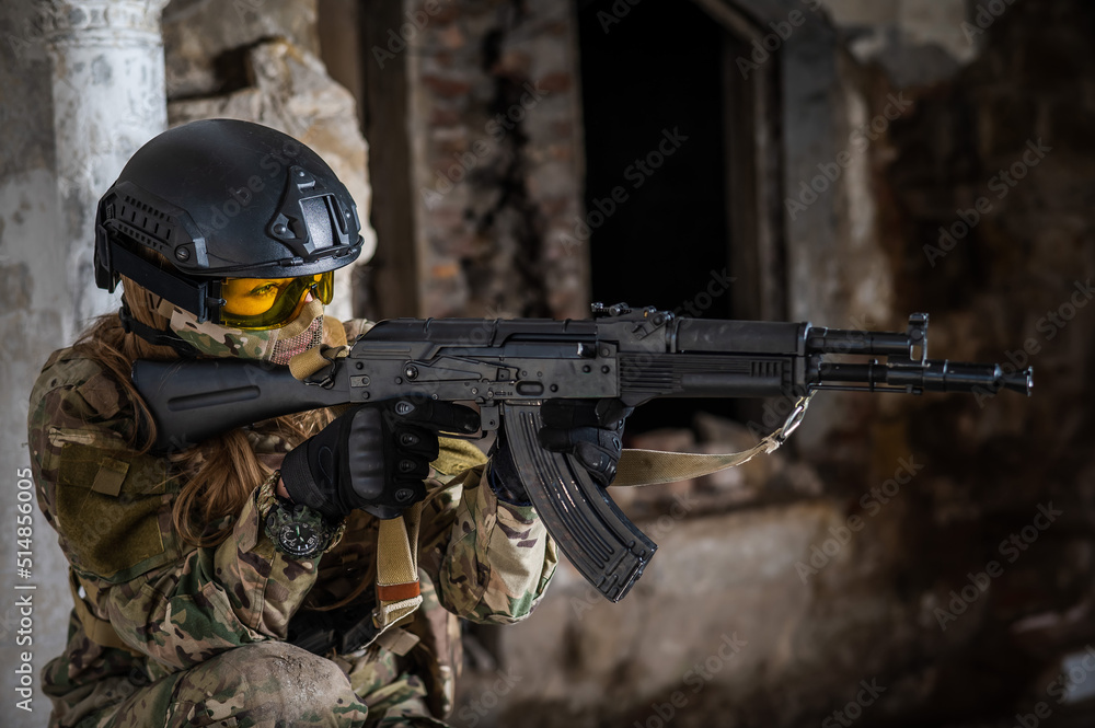 Fototapeta premium Portrait of a woman in a helmet and goggles with a machine gun in her hands. A female soldier in a camouflage uniform holds a weapon.