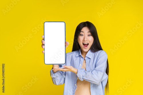 Super excited young asian girl with smartphone in hand showing white screen, mobile app, web service advertisement and excited looking at camera isolated on yellow background