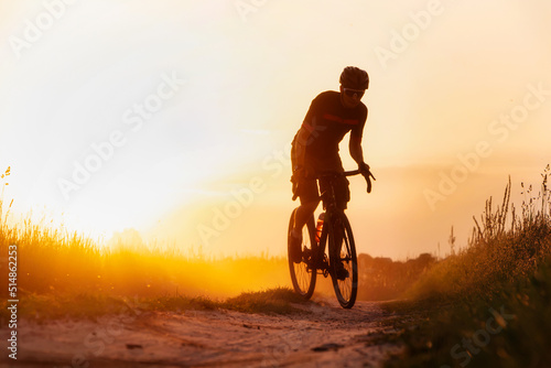 Silhouette young of cyclist on a gravel bike riding on a dust trail at sunset.