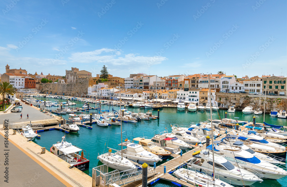 The old port harbor of the historic city of Ciutadella de Menorca, Spain, with fishing boats in the marina and the shops and cafes in view under the walled medieval city.