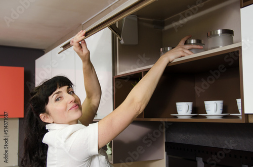 The smiling woman pulls a glass jar from a hinged cupboard, holding the raised door with her other hand.