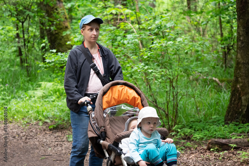 Young woman in dark jacket, jeans and a cap with headphones walks with her two sons along a forest road