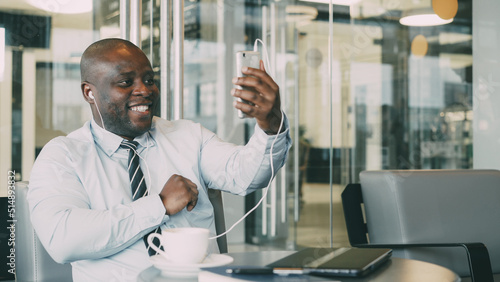 Portrait of African American businessman in formal clothes smiling and talking to his family through smartphone webcam in a modern cafe. He waving his hand happily having earbuds in his ears