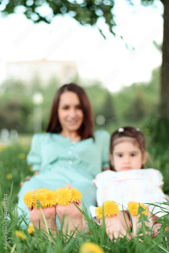 Blurry mother and daughter sitting on the grass in the park on a summer day.In the foreground are bare feet with dandelion flowers between the toes.Summer,closeness to nature,simple slow living. Blurry mother and daughter sitting on the grass in the park on a summer day.In the foreground are bare feet with dandelion flowers between the toes.Summer,closeness to nature,simple slow living.