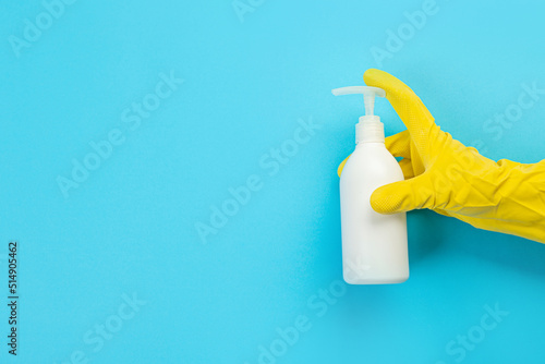 A hand in a yellow glove holds a white bottle of detergent on a blue background. Banner with copy space. Chemical cleaners, household chemicals, brushes and collage supplies. Cleaning concept.