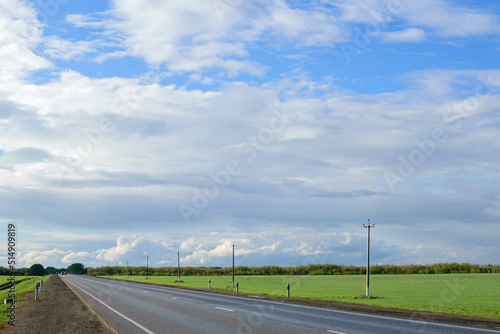 Long asphalt road between green fields under blue cloudy sky on a sunny summer day, summer road landscape
