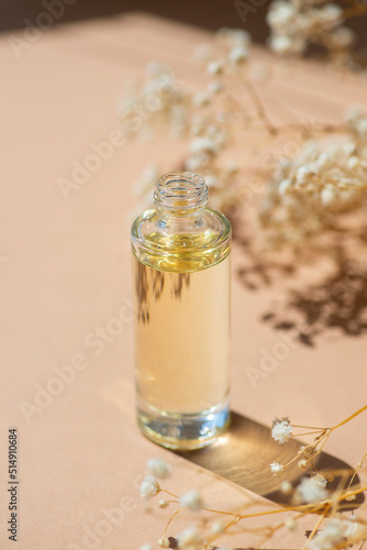 Open transparent bottle without cap with serum or essential oil with beautiful flowers in soft focus. Beige background with daylight and beautiful shadows. Beauty concept for face and body care