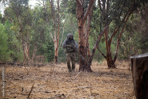 A soldier in a camouflage net walks with a sniper rifle in his hands. In the middle of the forest area, he is lying with a weapon in his hands. Professional airsoft player