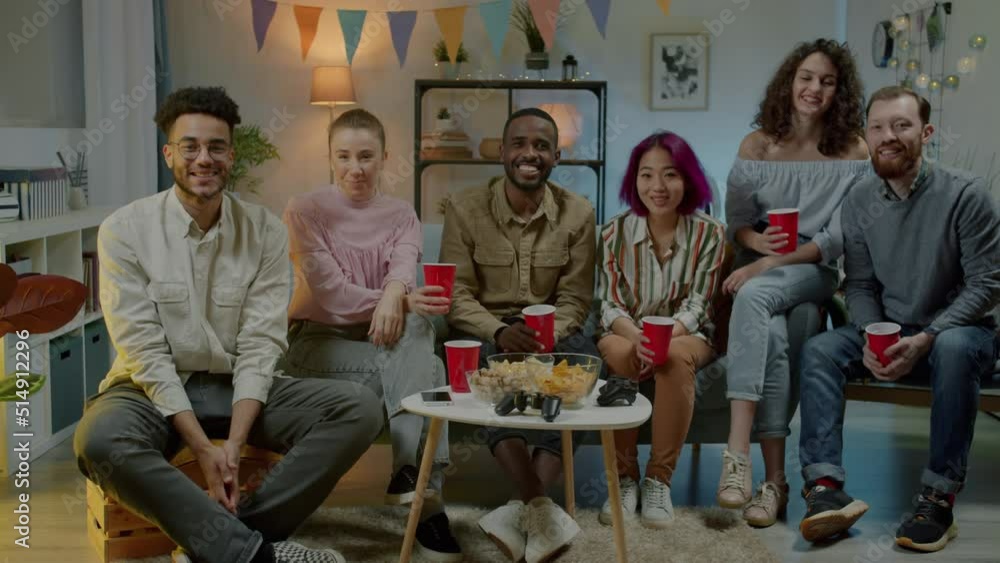 Slow motion portrait of young people raising cups with drinks and smiling looking at camera at party in dark apartment. Youth lifestyle and millennials concept. Slow motion portrait of young people raising cups with drinks and smiling looking at camera at party in dark apartment. Youth lifestyle and millennials concept.