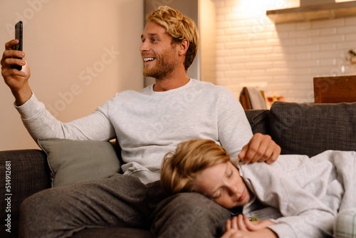 Ginger man using mobile phone while resting with his son on couch