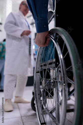 Closeup of older man hand using wheelchair in front of medical doctor for appointment in private hospital reception. Selective focus on senior man living with disability at medical clinic.