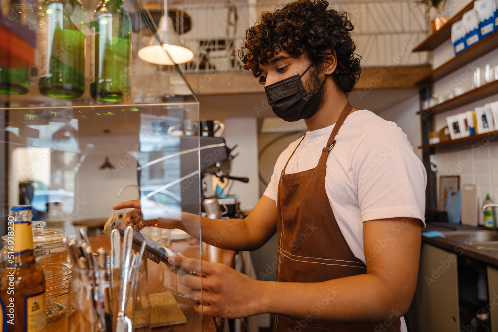 Fototapeta premium Young indian curly barista in face mask working with tablet