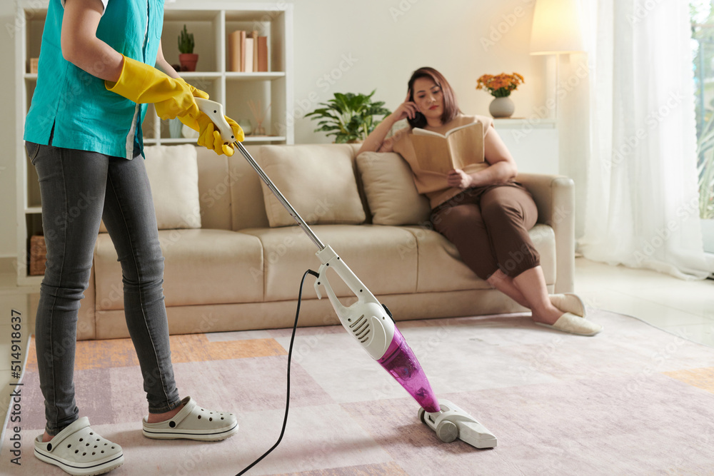 Close-up of housemaid in uniform using vaccum cleaner to clean the carpet in living room with owner sitting in background