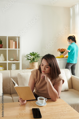 Asian young woman enjoying reading a book and cup of coffee during her leisure time while cleaner doing housework