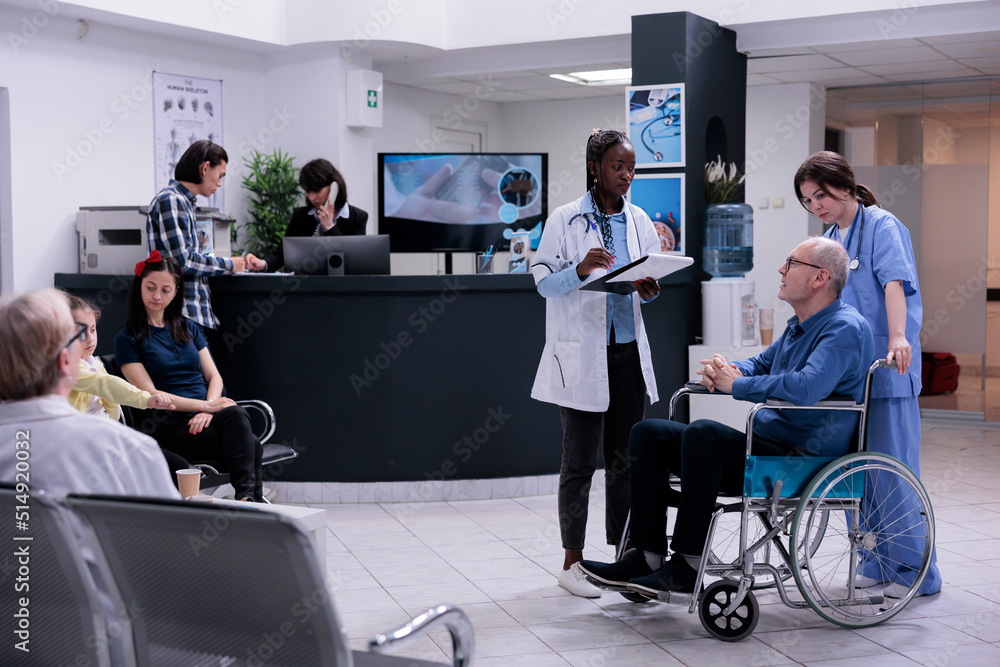 Retired person using wheelchair being helped by professional nurse talking with african american doctor for appointment. Older man living with disability in hospital reception with diverse people. Retired person using wheelchair being helped by professional nurse talking with african american doctor for appointment. Older man living with disability in hospital reception with diverse people.