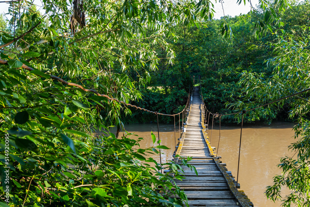Obraz premium Suspended wooden bridge to the other side of the river. Suspension bridge in a small village