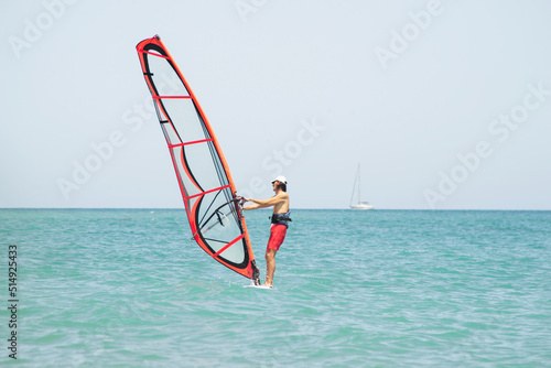 a man controls a sail on a surfboard in the sea against the background of the water and the sailboat in the distance