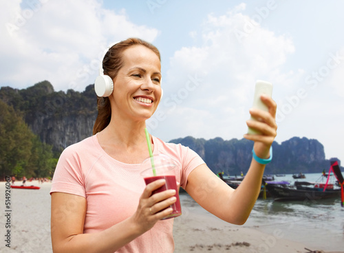 travel, tourism and vacation concept - woman in earphones with takeaway smoothie drink in plastic cup and smartphone listening to music over tropical beach background in french polynesia © Syda Productions