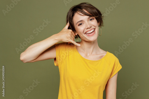 Young cheerful fun smiling happy woman she 20s wear yellow t-shirt doing phone gesture like says call me back isolated on plain olive green khaki background studio portrait. People lifestyle concept.