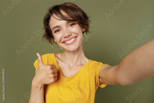 Close up young satisfied fun happy woman she 20s wear yellow t-shirt doing selfie shot pov on mobile cell phone show thumb up gesture isolated on plain olive green khaki background studio portrait