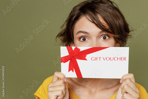 Young caucasian woman she 20s wear yellow t-shirt hold cover mouth with gift certificate coupon voucher card for store isolated on plain olive green khaki background studio. People lifestyle concept.
