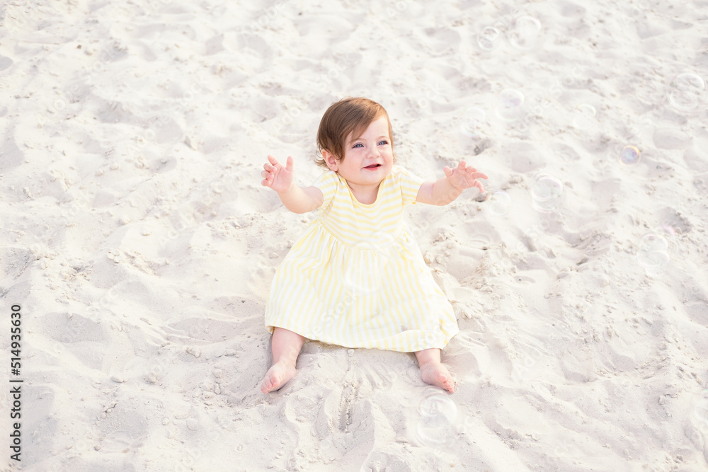 happy baby girl on beach on sand in striped yellow summer dress plays with soap bubbles on sunny day happy baby girl on beach on sand in striped yellow summer dress plays with soap bubbles on sunny day