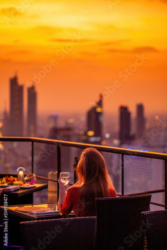 Young woman with glass of wine rest at luxury rooftop restaurant watching orange sky sunset. Female with cocktail drink at sky bar terrace looking at modern city skyline. Skyscrapers on background. photo