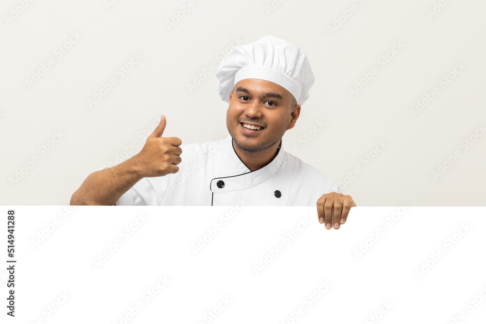 Happy Young handsome asian man chef in uniform looks out from behind an empty whiteboard. Cooking indian man hiding behind big blank billboard for advertising text menu in kitchen and restaurant. Happy Young handsome asian man chef in uniform looks out from behind an empty whiteboard. Cooking indian man hiding behind big blank billboard for advertising text menu in kitchen and restaurant.