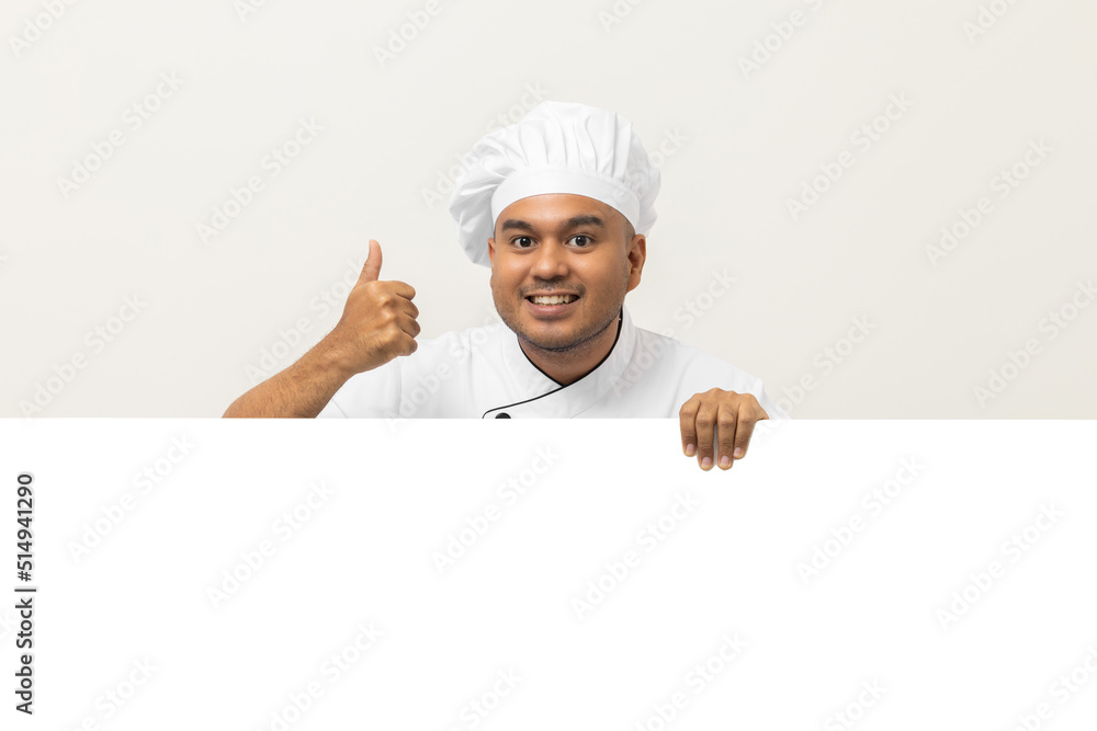 Happy Young handsome asian man chef in uniform looks out from behind an empty whiteboard. Cooking indian man hiding behind big blank billboard for advertising text menu in kitchen and restaurant. Happy Young handsome asian man chef in uniform looks out from behind an empty whiteboard. Cooking indian man hiding behind big blank billboard for advertising text menu in kitchen and restaurant.