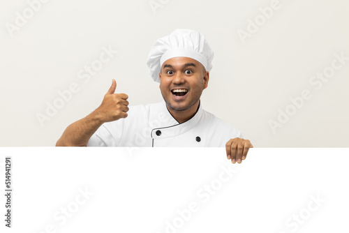 Happy Young handsome asian man chef in uniform looks out from behind an empty whiteboard. Cooking indian man hiding behind big blank billboard for advertising text menu in kitchen and restaurant.