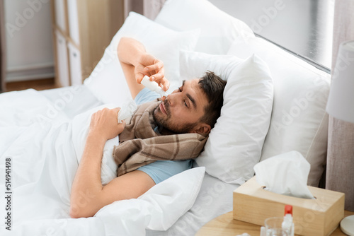people and health problem concept - unhappy sick man spraying his nose with nasal spray lying in bed at home