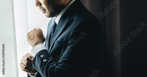 Confident businessman buttoning or adjust classic blue suit near window in hotel room at the morning. Handsome man wearing a nice suit on wedding day.