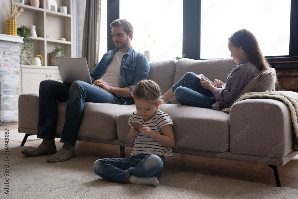 Young family of three holding and using different electronic devices while sitting on sofa in the living room