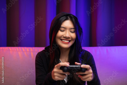 Playing video game. Young asian pretty woman sitting on sofa holding joystick in living room. Happy female Professional Streamer chinese wearing hoodie playing game online in dark room neon light.