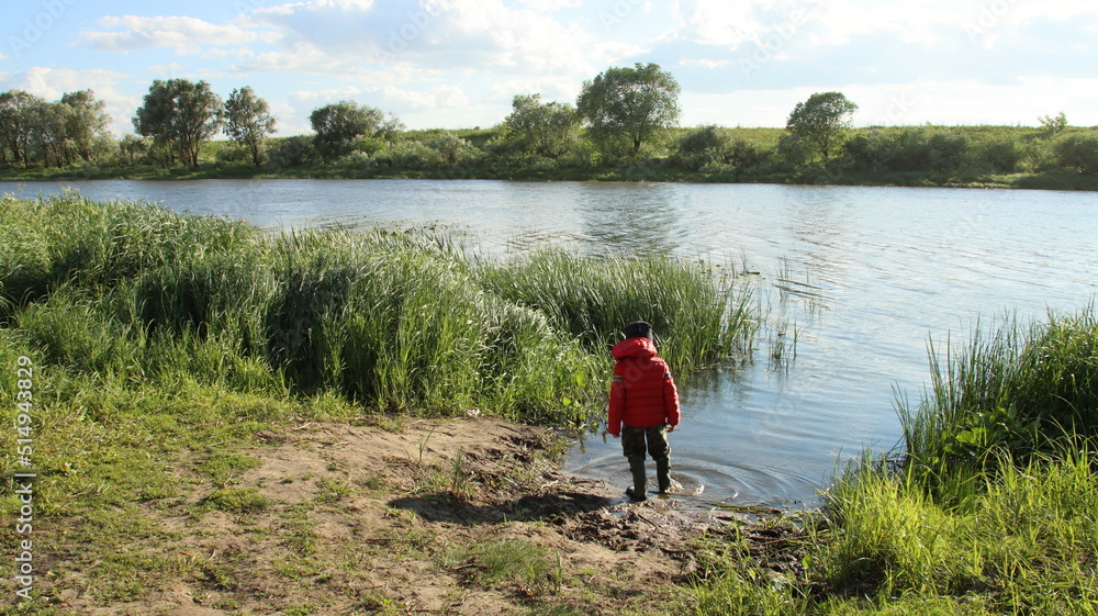 A boy in a red jacket and a blue hat stands on the swampy bank of the river