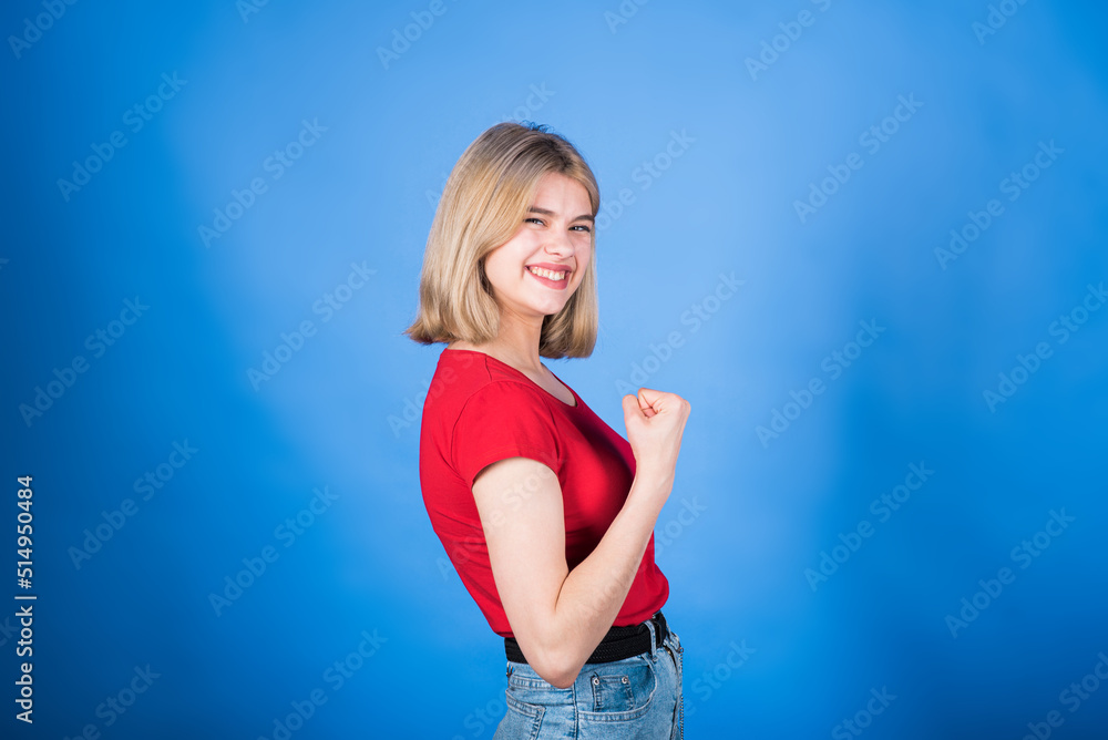 Young and attractive caucasian blonde girl in casual clothes clenching her fist and showing a winner gesture isolated on a blue studio background. Young and attractive caucasian blonde girl in casual clothes clenching her fist and showing a winner gesture isolated on a blue studio background.